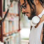 Confidence, Assertiveness - Confident adult Hispanic male in sunglasses and casual clothes with backpack and headphones reading book near bookshelf while making choice in stall on street