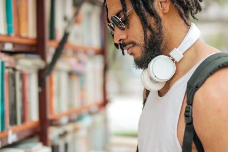Confidence, Assertiveness - Confident adult Hispanic male in sunglasses and casual clothes with backpack and headphones reading book near bookshelf while making choice in stall on street
