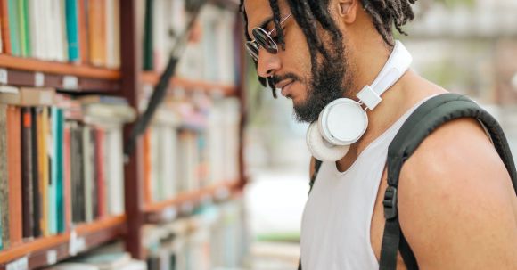 Confidence, Assertiveness - Confident adult Hispanic male in sunglasses and casual clothes with backpack and headphones reading book near bookshelf while making choice in stall on street