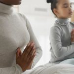 Mindfulness Practice - Full body of tranquil young barefooted African American woman in casual clothes sitting on carpet in Padmasana pose with closed eyes while meditating with adorable little daughter at home