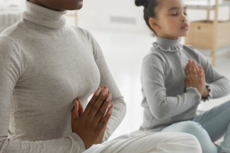 Mindfulness Practice - Full body of tranquil young barefooted African American woman in casual clothes sitting on carpet in Padmasana pose with closed eyes while meditating with adorable little daughter at home