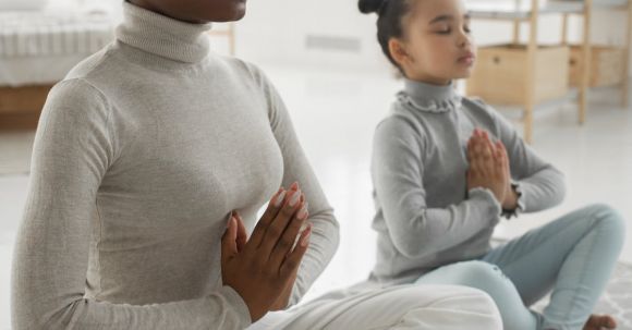 Mindfulness Practice - Full body of tranquil young barefooted African American woman in casual clothes sitting on carpet in Padmasana pose with closed eyes while meditating with adorable little daughter at home